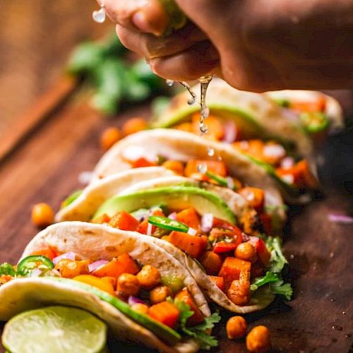 The image shows a hand squeezing lime over a row of fresh tacos filled with vegetables and chickpeas on a wooden board, garnished with lime slices.